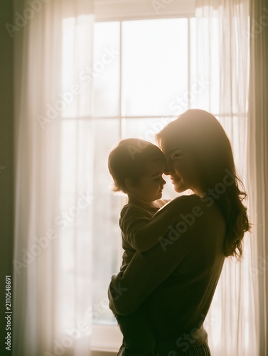 A mother and child share an intimate, tender moment as the mother holds her young child close by a bright sunlit window with sheer curtains. Soft natural light creates a warm, loving atmosphere.