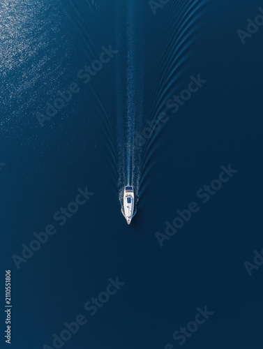 Top-down aerial view of a white sailboat gliding across calm, deep blue cobalt sea with gentle wake trails. Serene maritime scene capturing tranquility and natural beauty.
