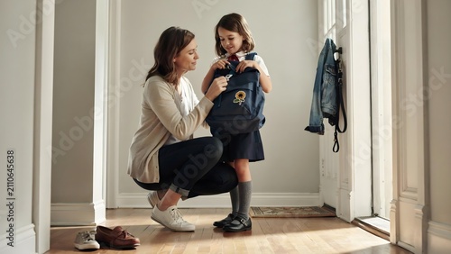 A caring mother helping her young daughter put on her school backpack in the hallway before school, preparing for the day ahead