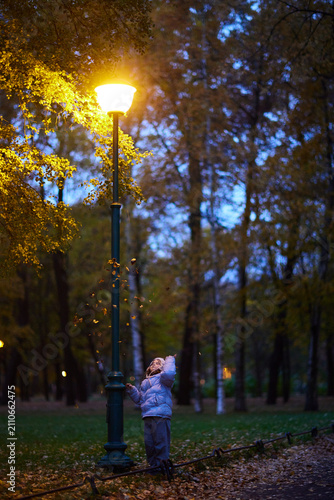 Child plays with leaves near a street lamp in a park during the evening