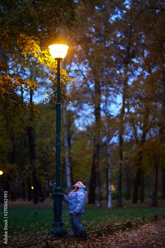 Child stands near a street lamp in a park during dusk while looking up at the light and enjoying falling leaves