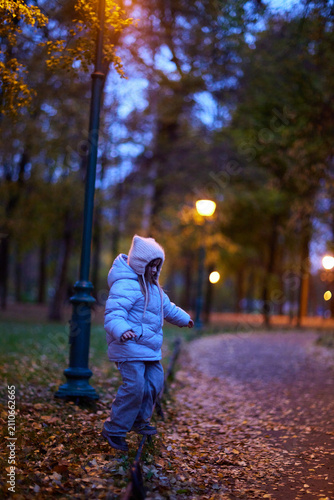 Child walks along a pathway in a park during evening with street lights shining and fallen leaves on the ground