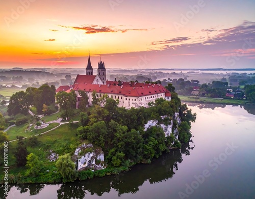 Aerial view captures a serene dawn over a historical European structure on a cliffside, beside a calm river with a hazy ambiance