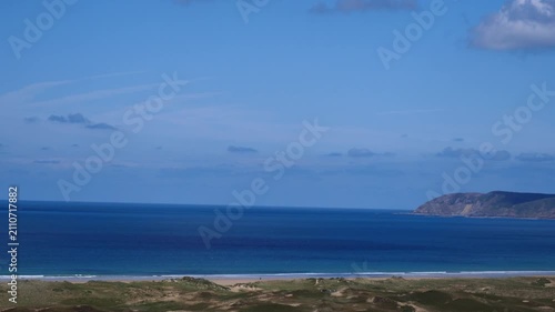 Calm ocean meets sandy shore under blue sky. Dunes and greenery frame the coastline. Distant hills and scattered clouds complete the peaceful scene