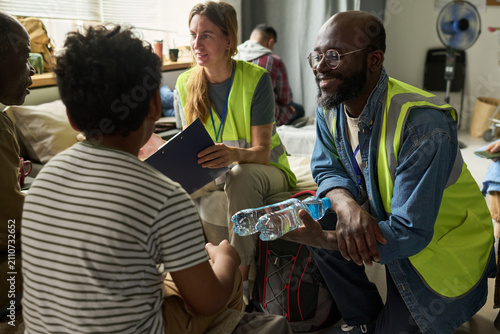 Canvastavla Medium shot of diverse aid workers distributing water and coordinating support with displaced adults in shelter, representing humanitarian relief