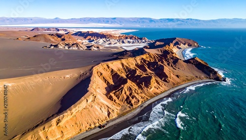 Aerial view of coastal formations where desert meets ocean. Rugged terrain contrasted with smooth, turquoise water, with mountains in the distance