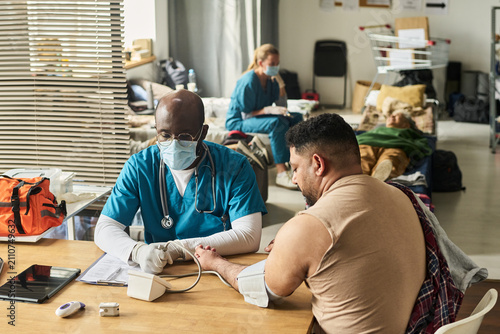 Medium shot of Black male doctor checking middle aged man blood pressure in temporary shelter clinic, providing urgent care. Useful for healthcare access, crisis response, outreach campaigns