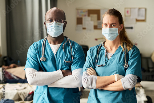 Obraz na plátně Portrait of African American male doctor and Caucasian female nurse standing with crossed arms, wearing masks in clinic