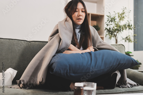 sick asian young woman sneezing into tissue paper while covered with a blanket and sitting on sofa in the living room