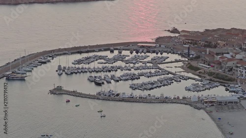 Drone view above the yacht-filled marina of Isola Rossa in Sardinia, Italy, surrounded by breakwaters, seaside buildings and calm Mediterranean water at sunset.