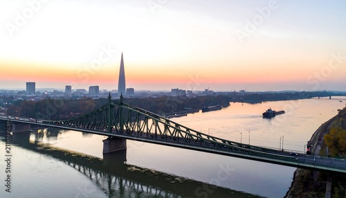 Aerial view captures a wide bridge spanning a vast river, a distant city skyline with a tall tower, and an autumn sunset