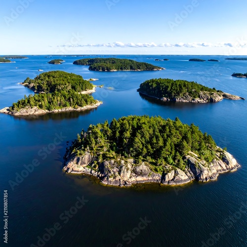Aerial view captures numerous green, forested islands scattered across deep blue waters under a clear, bright sky. Sunlight highlights details