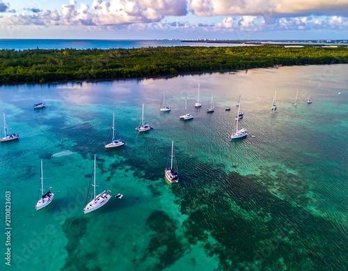 Aerial view captures sailboats anchored in crystal clear turquoise water, near lush green vegetation, under a partially clouded sky