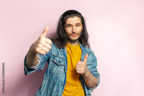 Young happy man with long hair showing thumbs up. Isolated on pastel pink background.