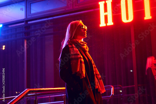Schilderij op canvas Woman with windswept hair standing in profile beneath glowing marquee sign at ni