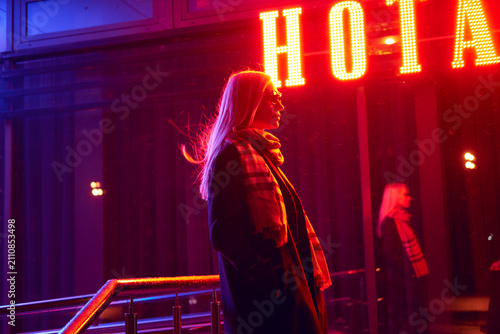 Schilderij op canvas Woman with windswept hair standing in profile beneath glowing marquee sign at ni