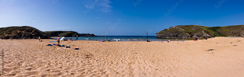Fototapeta premium Wide panoramic view of a sunny sandy beach with rocky cliffs, calm blue sea and people relaxing, swimming and surfing on a warm summer holiday at the coast.