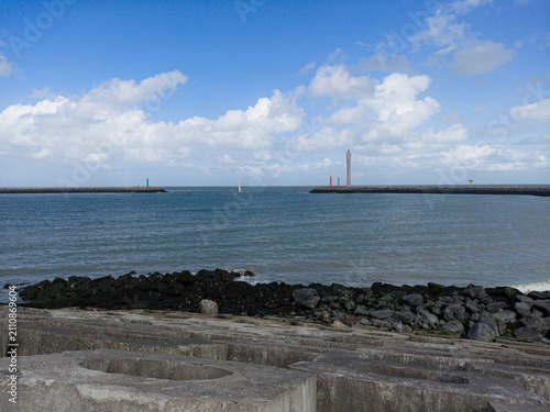 A calm harbor entrance with concrete breakwater and rocky shore, distant lighthouses and a solitary sailboat on the blue sea beneath a bright sky filled with scattered white clouds.