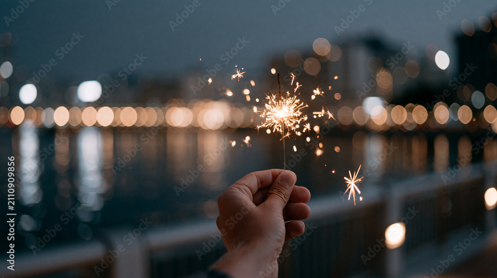 Fototapeta premium Hand holding a sparkler at dusk near the water, capturing sparkling moments of joy Generative AI