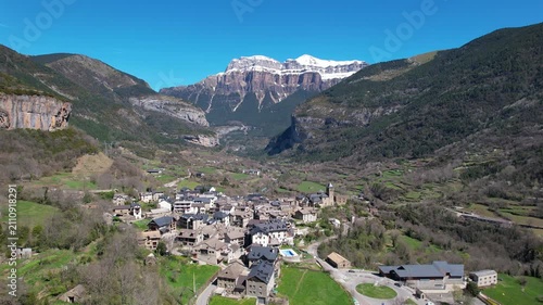 Forward reveal aerial shot of Torla-Ordesa village and covered with snow mountains of Ordesa y Monte Perdido National Park on sunny spring day. Huesca, Aragon, Spain.