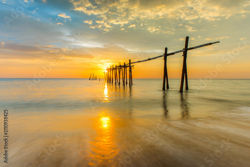 Fotografie The remains of an old wooden bridge, with only the structural pillars left, lie by the sea in the evening as the sun is setting