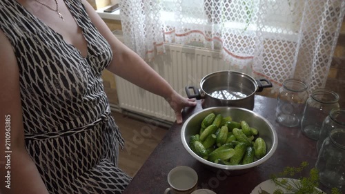 Woman preparing cucumbers for canning in the kitchen, Woman standing near table with fresh cucumbers and glass jars for preservation