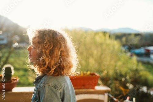 Fotografía Teen girl with curly hair stands on a balcony, enjoying a sunny day