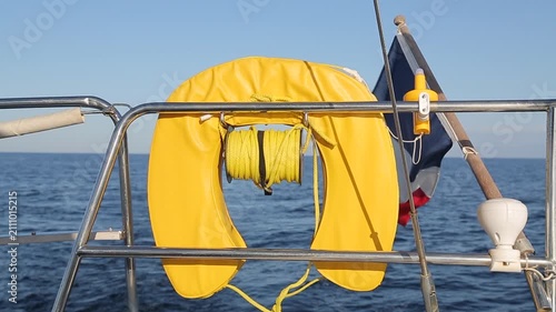 Close-up shot from the stern of a sailing yacht looking backward at a yellow horseshoe lifebuoy and rescue rope reel attached to the stainless steel pushpit railing.  
