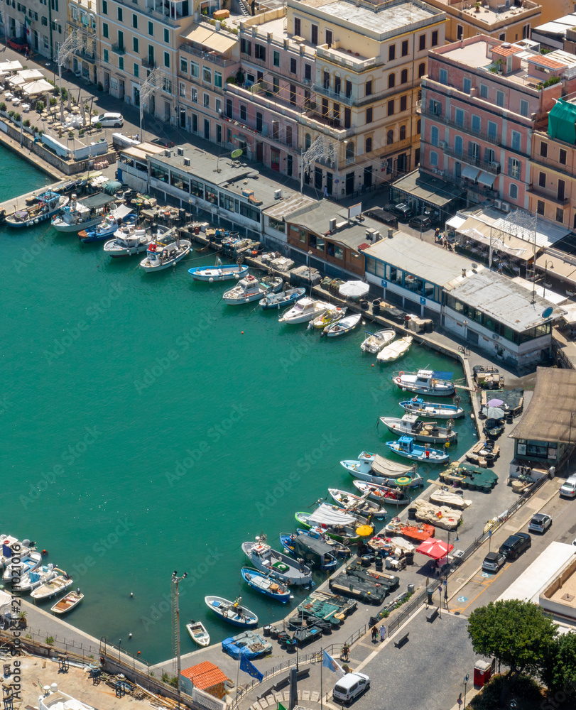 Fototapeta premium Aerial top view of a scenic coastal harbor featuring boats docked along a curved stone promenade lined with charming buildings and vibrant turquoise sea water. It's Anzio, a coastal town in Italy.
