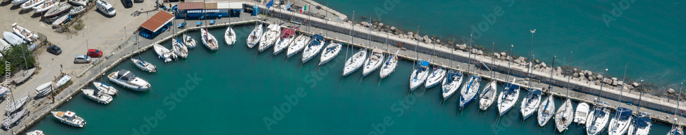 Obraz premium Aerial panoramic view of a picturesque marina filled with numerous white boats and yachts docked along a stone pier, surrounded by clear turquoise sea water on a sunny bright day.