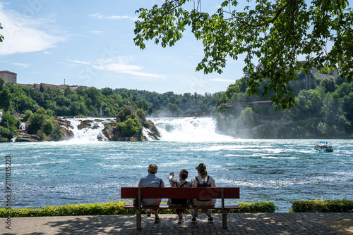 Group of elderly people resting on bench and admiring view of Rhine Falls waterfall in summer, back view