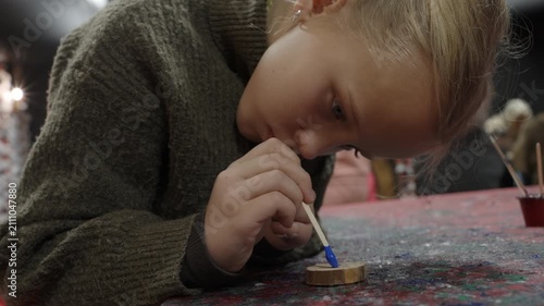 Focused blonde girl paints a small wooden slice with bright blue paint using a stick, concentrating on her art and craft project at a table