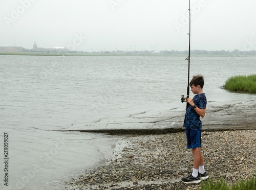 Canvas Print A young boy stands on a pebbled shore concentrating as he reels in a line at a calm, overcast riverbank, capturing quiet patience, outdoor recreation and a summertime hobby by the water