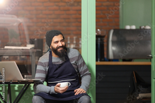 Full length portrait of a smiling male coffee shop owner or barista holding a coffee cup while sitting on a chair inside a modern cafe.