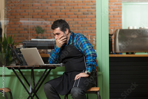A stressed and worried coffee shop owner or barista wearing an apron sitting at a table inside a cafe, looking at a laptop computer with concern, anxiety, or financial trouble.