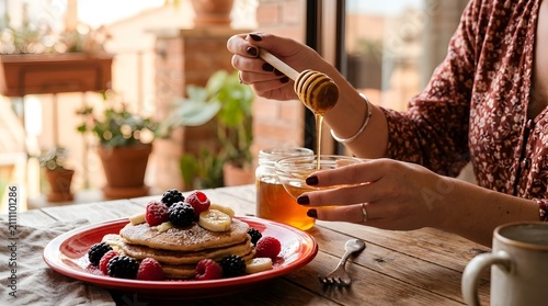 Woman Drizzling Honey on Pancakes with Berries and Bananas for Breakfast.