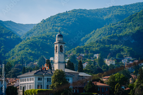 Photography Scenic view of Carate Urio with historic clock tower and lush green mountains in