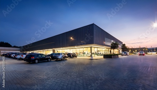 a wide view of a modern car dealership at dusk with cars lined up outside and lights illuminating the showroom