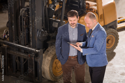 High angle view of two businessmen without hard hats standing in front of a forklift and looking at a digital tablet inside a warehouse.