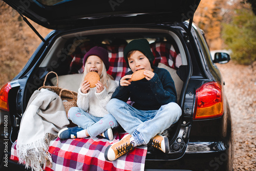 Cute children having picnic in car trunk sitting on checkered blanket in woods over autumn leaves. Fall season. Childhood.