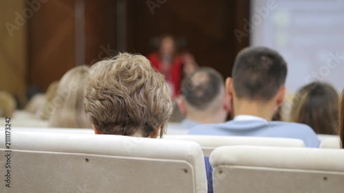 Audience members watch presentation from behind. People focus on speaker, showing interest. Scene shows conference or seminar setting. Ideal for business, education, or corporate events