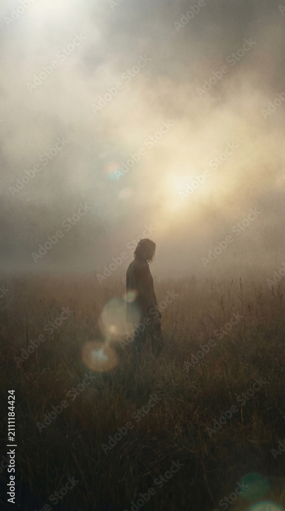 Fototapeta premium Silhouette Of A Lone Person Standing In A Grassy Field At Sunrise With Morning Mist And Lens Flare