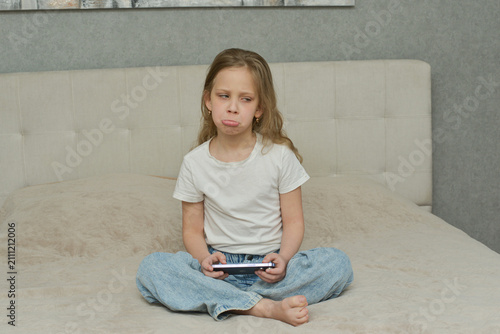 Unhappy Young Girl Sitting on Bed Holding Smartphone. A candid photograph of a young girl, approximately 7-10 years old. She has long, wavy blonde hair 