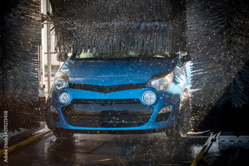 Фотография Straight-on close-up of a blue hatchback's front end being scrubbed by black rotating car wash brushes, water and suds spraying across the headlights and grille