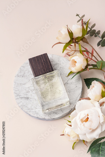 Glass perfume bottle on a round marble podium with floral decoration on a pastel beige background