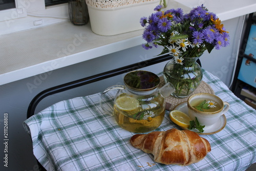 Herbal tea with lemon and a croissant placed on a checkered tablecloth, accompanied by a vase of wildflowers in a bright kitchen near a window