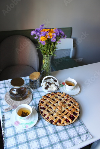 A fruit pie sits on a checkered tablecloth alongside tea cups and a teapot, with a vase of colorful flowers in a cozy dining space