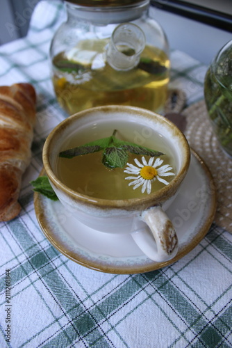 Herbal tea in a decorative cup with mint leaves and a daisy flower, accompanied by a croissant on a checkered tablecloth in a cozy kitchen setting