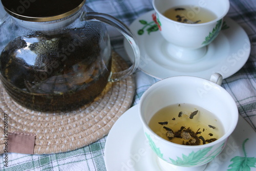 Glass teapot with brewed herbal tea beside two white cups on a checkered tablecloth, showcasing tea leaves floating in the cups and a woven coaster underneath