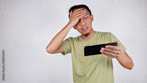 Canvastavla Young Indonesian man in green clothes touching his forehead while playing game i